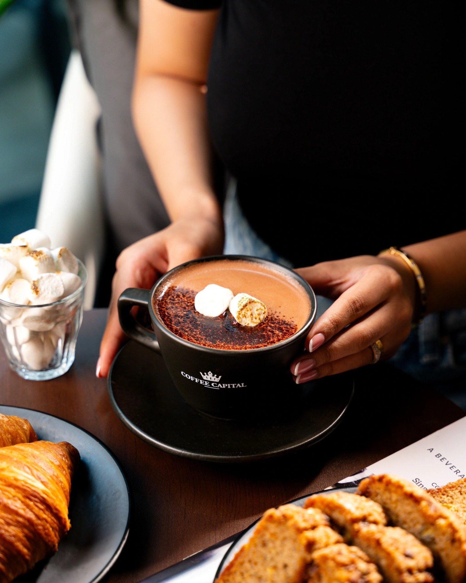 A Coffee Capital branded hot chocolate with marshmallows, served alongside croissant and biscotti